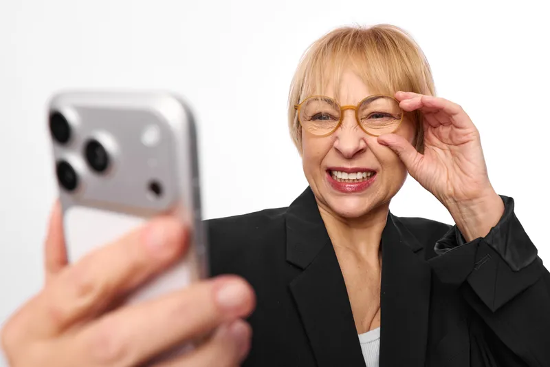 Senior woman with glasses smiling smartphone portrait, hand near cheek, studio background, cheerful mature expression, shallow depth of field, soft light