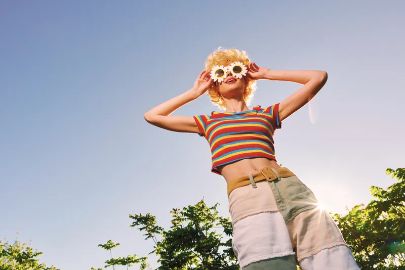 Woman lifestyle outdoor scene with retro vintage fashion, wearing striped crop top and patchwork shorts, flower sunglasses, exuding a sunny joyful mood and playful energy