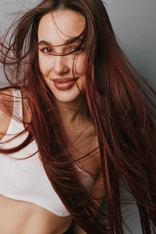 Woman with long hair smiling and wearing white top in studio. Portrait of happy female with natural makeup and flowing hair on gray background for beauty and fashion.