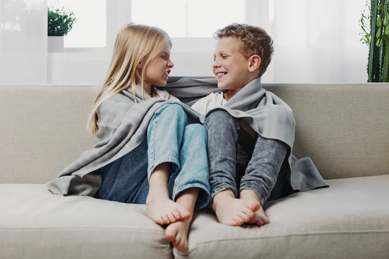 Two children wrapped in grey blankets sit barefoot on a sofa facing each other, smiling and enjoying a cozy moment in a bright living room with plants in the background.
