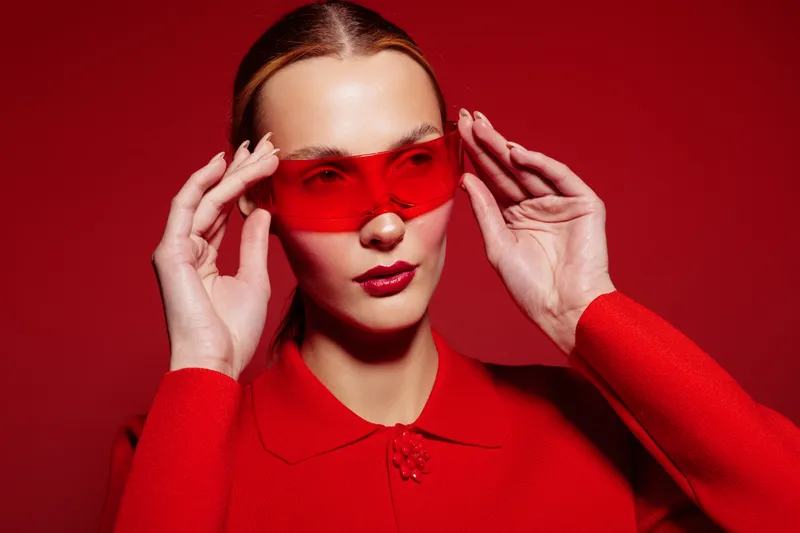 Stylish woman in red sweater adjusts red translucent glasses on red background. Fashion portrait with bold color contrast and modern accessories. Concept of style, confidence, and elegance.