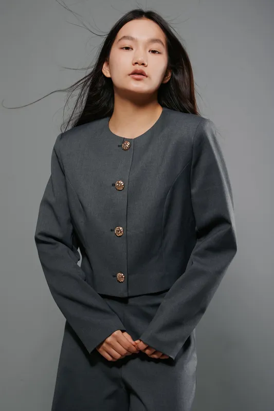 Young woman in gray suit with long hair standing with hands clasped in front against a plain background, expressionless face and natural look for fashion portrait.