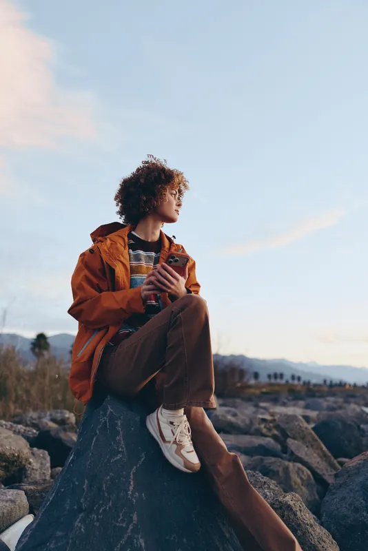 Woman outdoors lifestyle, casual jacket, sits on rock at sunset, curly hair, sneakers on feet, relaxed pose, blue sky with clouds, scenic nature travel mood.