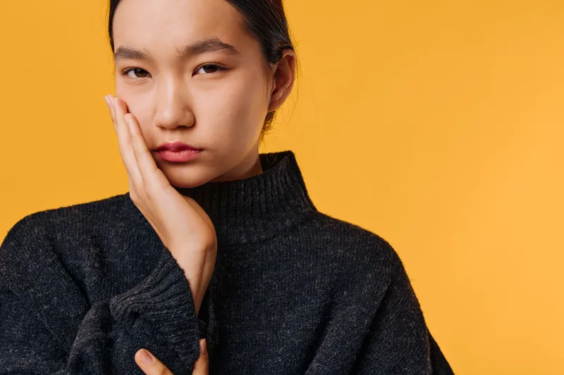 Young woman with hand on face looking serious and thoughtful wearing dark sweater on orange background for emotion and contemplation concept.