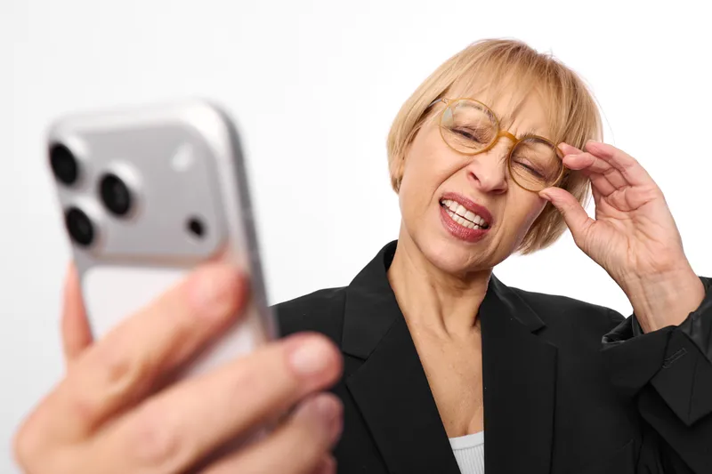 Elderly woman, smiling, adjusting glasses while holding phone, indoors, business attire, cheerful call moment, soft light, white background, focus on face, closeup