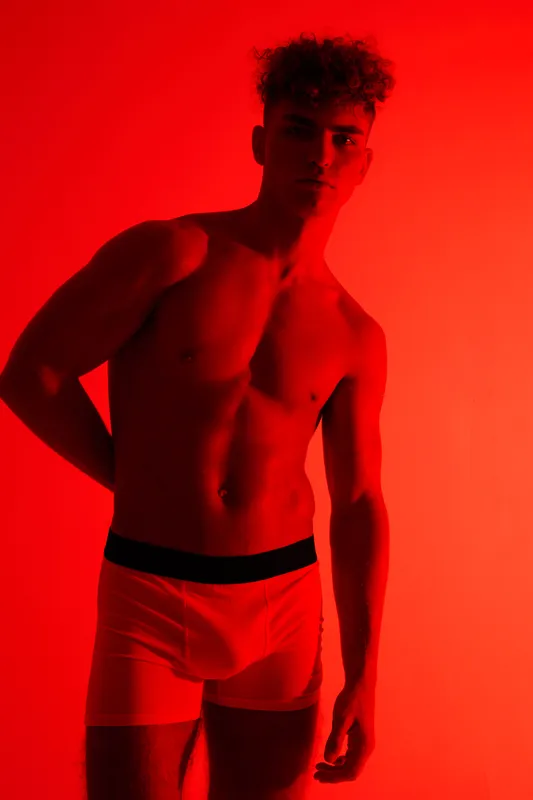 Young man wearing white underwear posing against red background with casual confident expression. Muscular torso, curly hair and dramatic lighting for artistic portrait session.