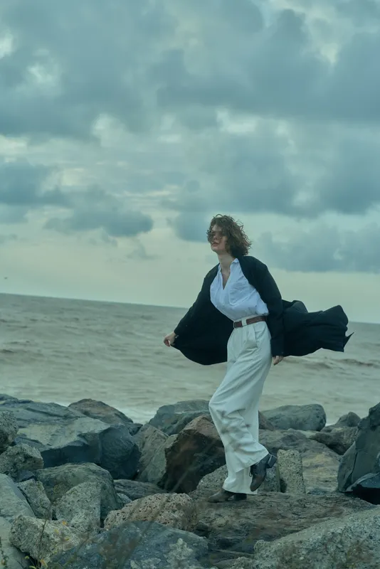 Woman in white suit and blazer walks along windy rocky shore, fashion movement by the sea, ocean backdrop and dramatic coastal scene