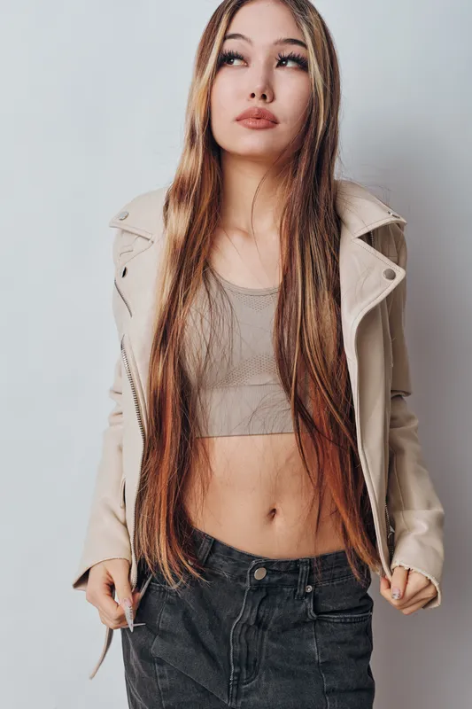Young woman with long hair wearing beige jacket and cropped top posing against gray background. Casual style, fashion, beauty and confident mood with natural makeup and relaxed posture.