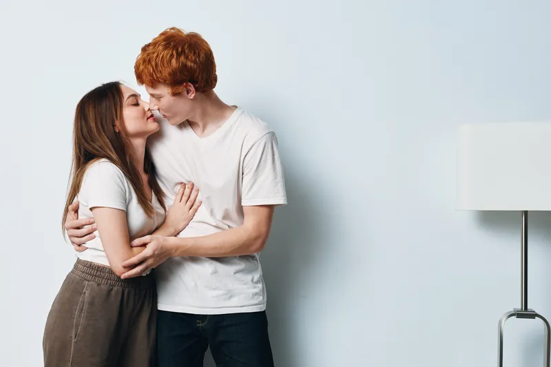 Couple kissing and hugging indoors, young man and woman in casual clothing show affection in minimalist room with neutral wall and floor lamp side light.