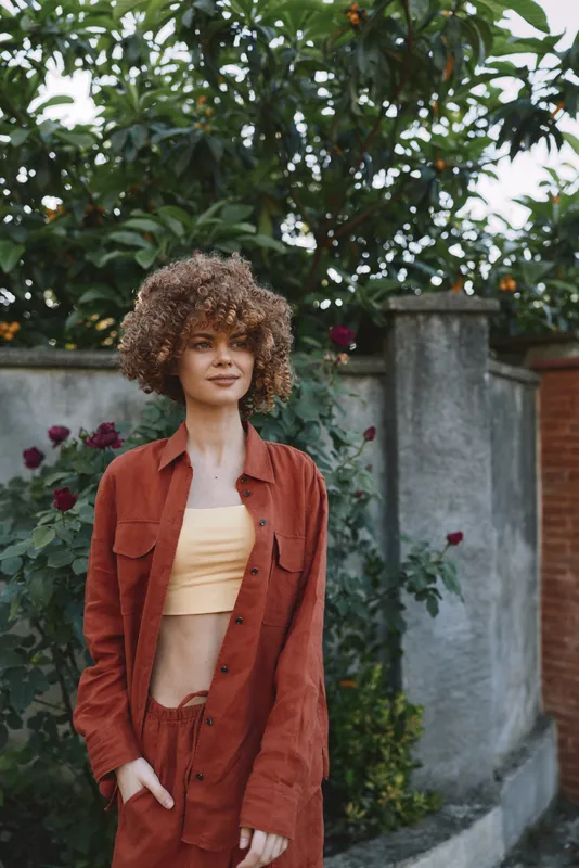 A young woman with curly hair stands in a sunny garden, wearing a rust orange jacket over a pale crop top, posing confidently with hands in pockets