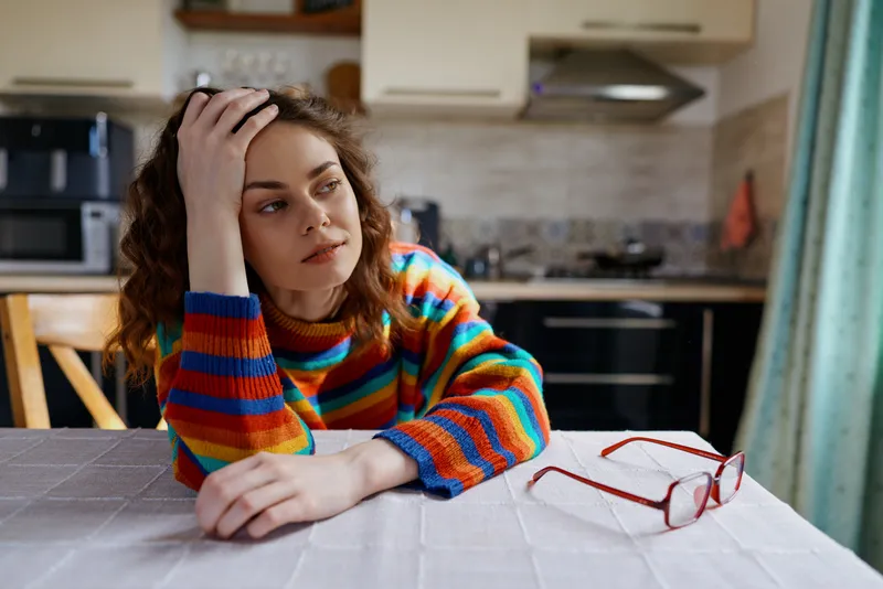 Thoughtful young woman with curly hair rests her head on her hand at a kitchen table, wearing a bright striped sweater, with red glasses on the table, a calm reflective mood in home setting.