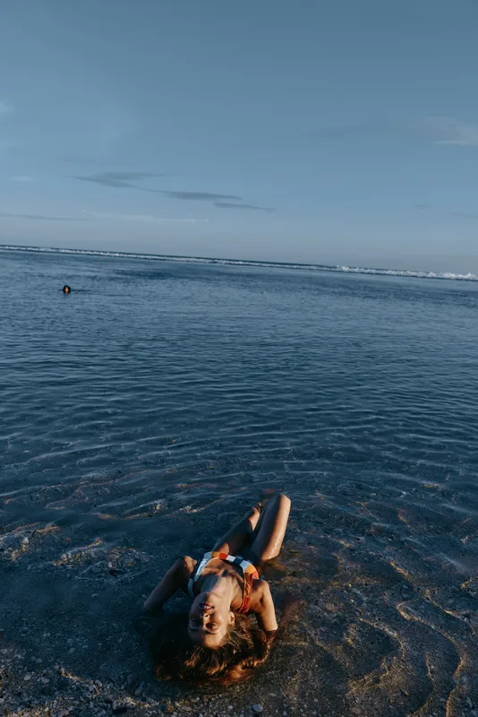 Beach ocean shoreline, woman in shallow water with arms up, dusk sky and soft clouds, summer relaxation, calm nature travel scene, blue evening light, quiet seascape