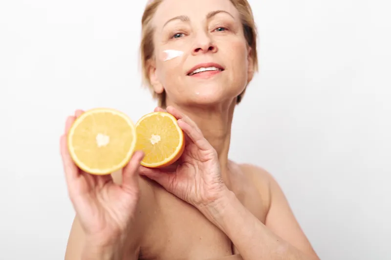 Mature woman with natural skin applies citrus face cream holding two halves of fresh orange, isolated on white background. Concept of skincare, healthy lifestyle, and natural beauty care.