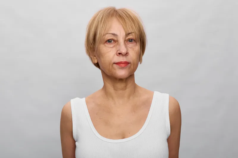 Elderly woman portrait studio background gray mature face expression, calm neutral look, short hair, sleeveless white clothing, closeup headshot, minimal light, copyspace