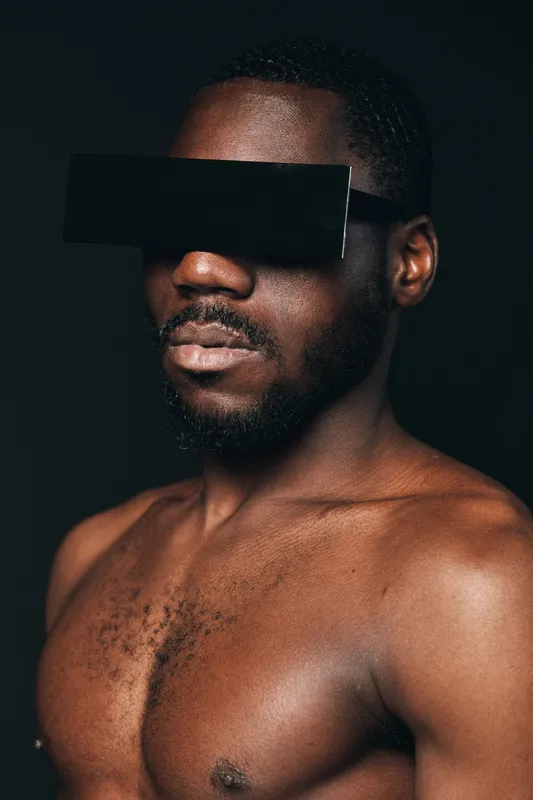 African man with dark skin and beard wearing futuristic black visor in creative conceptual visual style on black background. Portrait of confident male model with bare chest and modern accessory.