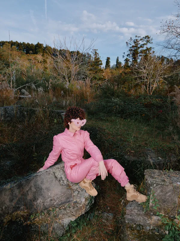 Woman in pink outfit sitting on a rock in an outdoor setting, surrounded by autumn foliage and trees, capturing a calm moment amid nature and rugged terrain, with soft daylight and tranquil