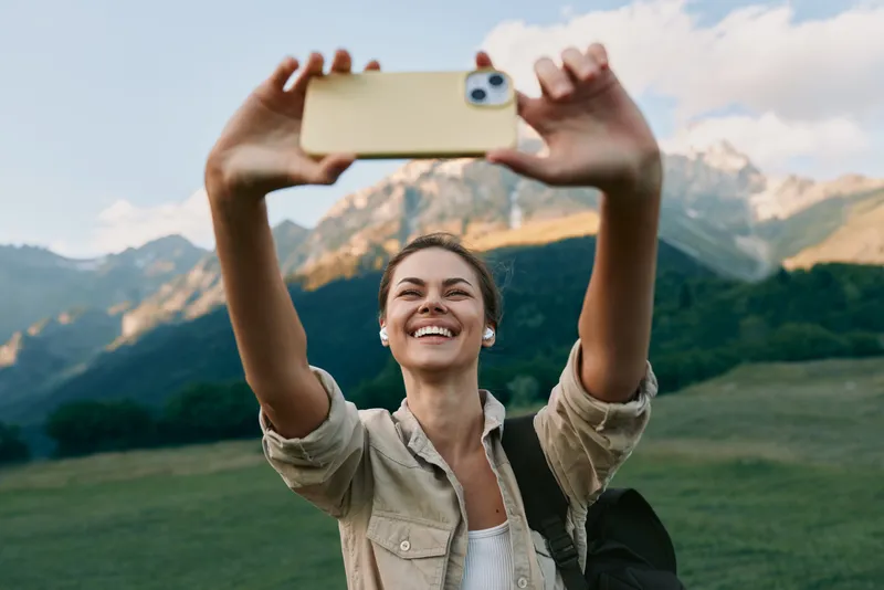 Young woman with a backpack takes a selfie with a smartphone in a mountainous landscape while smiling joyfully during a clear day.