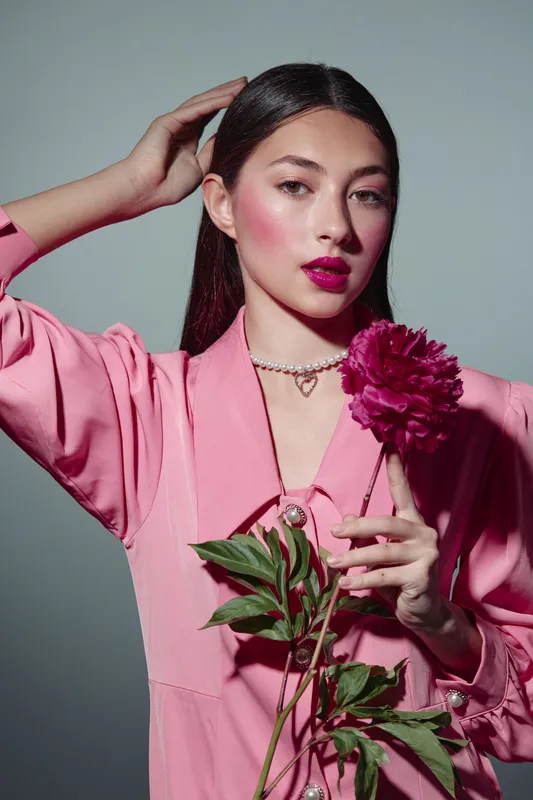 Elegant woman in pink blouse holds a dark magenta peony flower while posing thoughtfully in a studio. Her makeup is creative, featuring bold fuchsia lips and flushed cheeks. Beauty and fashion