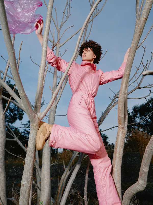 Person in a pink jumpsuit climbs a leafless tree outdoors holding a flowing scarf, an artistic fashion moment against a clear sky emphasizing movement, balance and bold color.