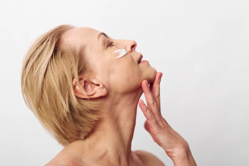 Mature woman with short blonde hair applies cream under eye, touching face gently, isolated on white background. Concept of skincare, aging, self-care, and beauty routine.