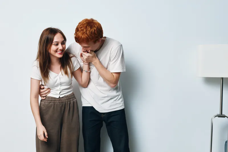 Young couple laughing and showing affection in casual clothes standing near white wall with lamp inside room, happy together and enjoying moment.
