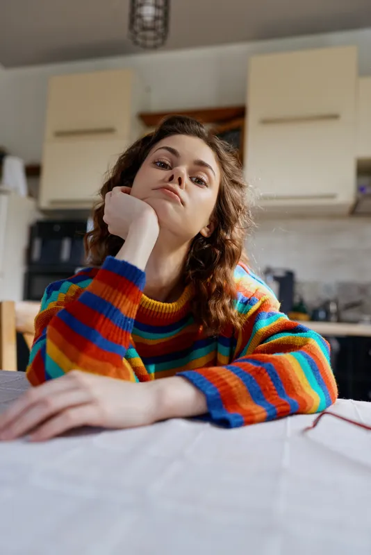 A young woman sits at a kitchen table in a colorful striped sweater, resting her chin on her hand and gazing thoughtfully at the camera, conveying a moment of contemplation.