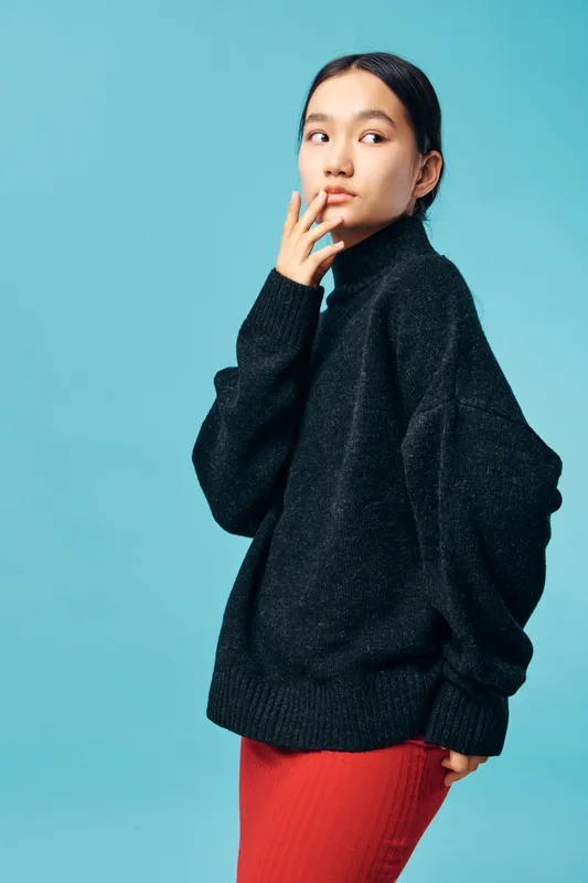 Young woman in black oversized sweater and red skirt posing against blue background looking sideways with thoughtful expression. Casual fashion, portrait, studio, model, style.
