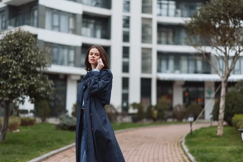 Young woman in a long dark coat walks thoughtfully along a paved path in an urban park surrounded by modern buildings and greenery on a cloudy day.