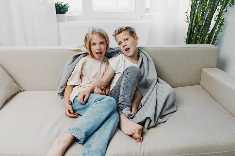 Two surprised children sitting on beige sofa wrapped in blanket, cozy home interior with natural light and plants. Concept of childhood, family, and casual moments.