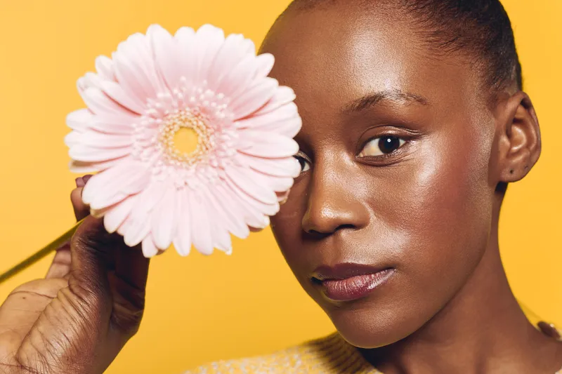 Close-up of a young black woman with dark skin holding a pink flower near her face against a yellow background. Concept of natural beauty, floral, and skincare wellness.