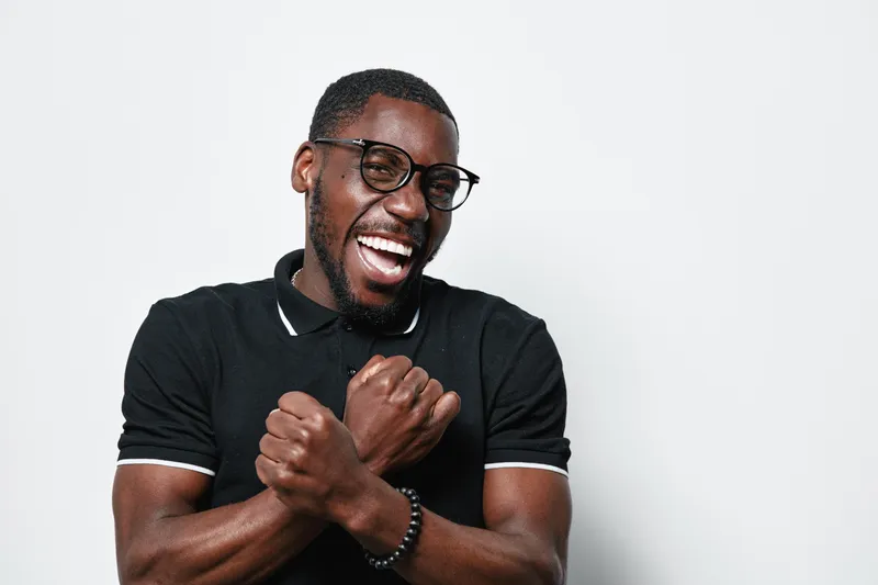 African American man with dark skin and glasses smiling and crossing arms in front of chest wearing black polo shirt on white background. Happy, confident and casual portrait.