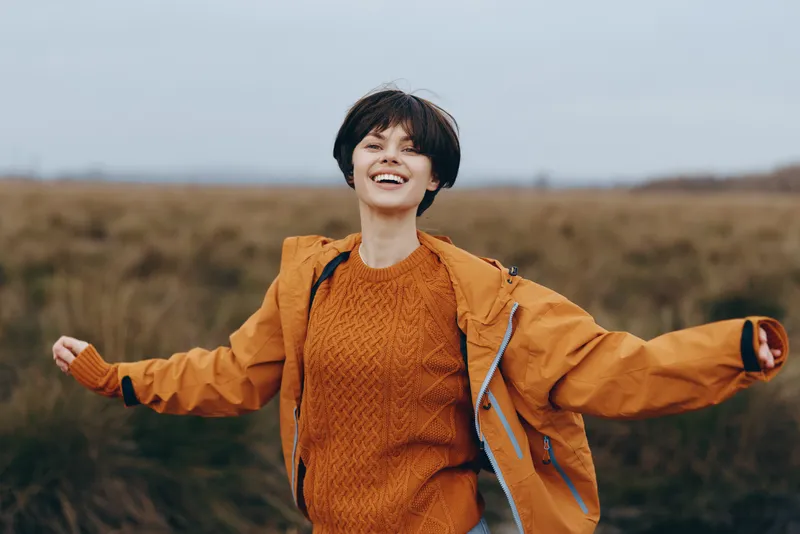 Woman hiking outdoors countryside, backpack jacket lifestyle travel moment, smiling woman in orange sweater and jacket, arms open running in open field, natural daylight