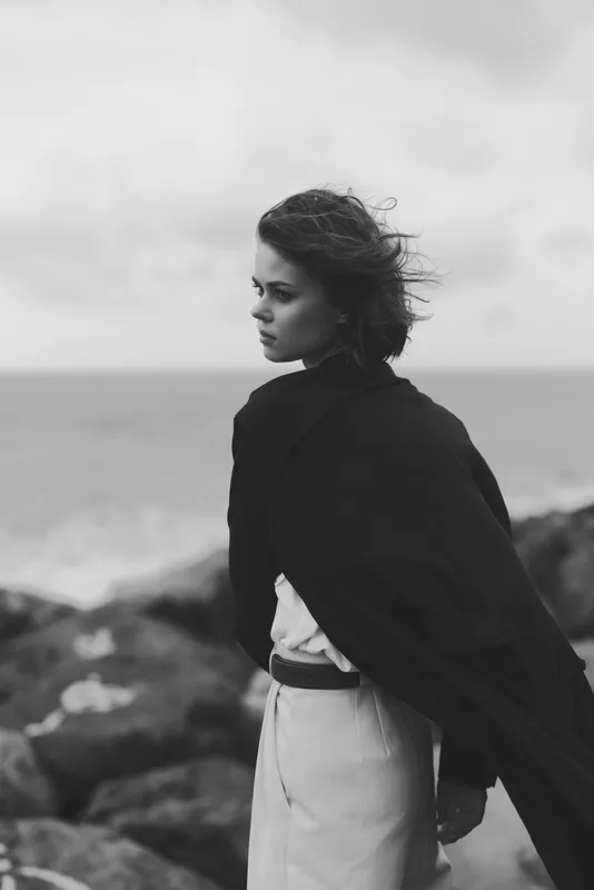 Woman, coat, shore, wind, cape, black and white mood, sea horizon, thoughtful pose on rocky beach, monochrome seascape with dramatic wind swept fabric