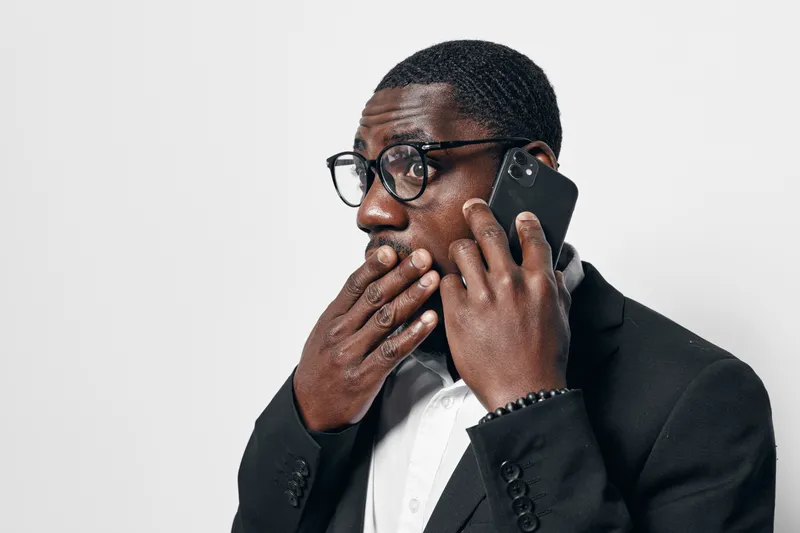 African American man with dark skin wearing glasses and black suit talking on smartphone with surprised expression covering mouth on white background.