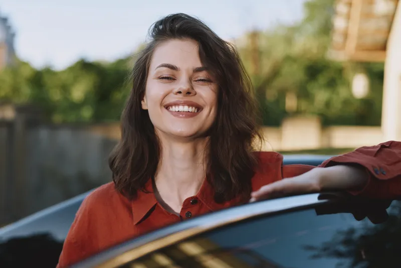 Cheerful woman outdoors, smiling with eyes closed, sunlit moment, casual red shirt, leaning on car, relaxed lifestyle scene in warm natural colors, with a soft breeze