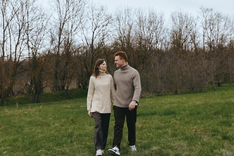 Young couple wearing knitted sweaters walks together on a green meadow in early spring with bare trees in the background, enjoying a calm and intimate moment outdoors.