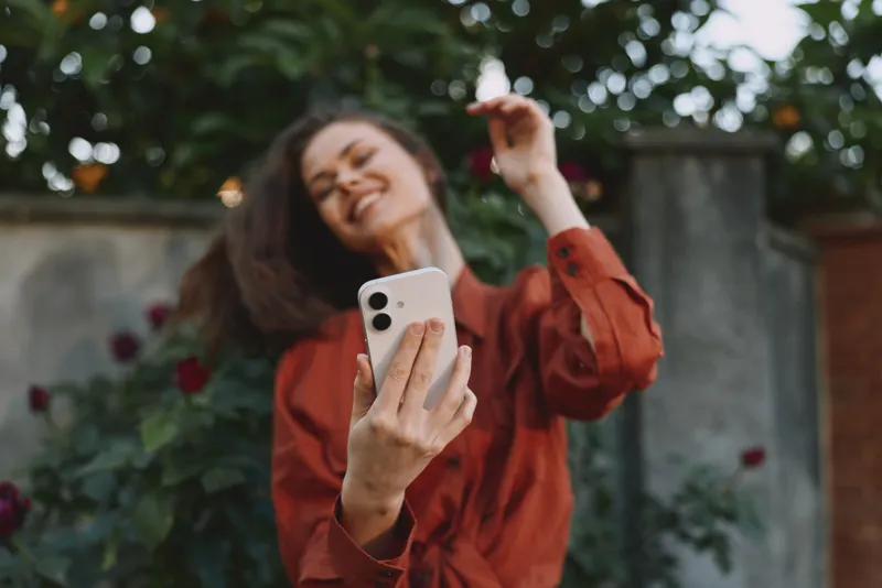 Young woman in a red blouse takes a cheerful selfie in a garden setting, capturing a carefree lifestyle moment with sunny ambiance and relaxed pose. Across a quiet background wall scene