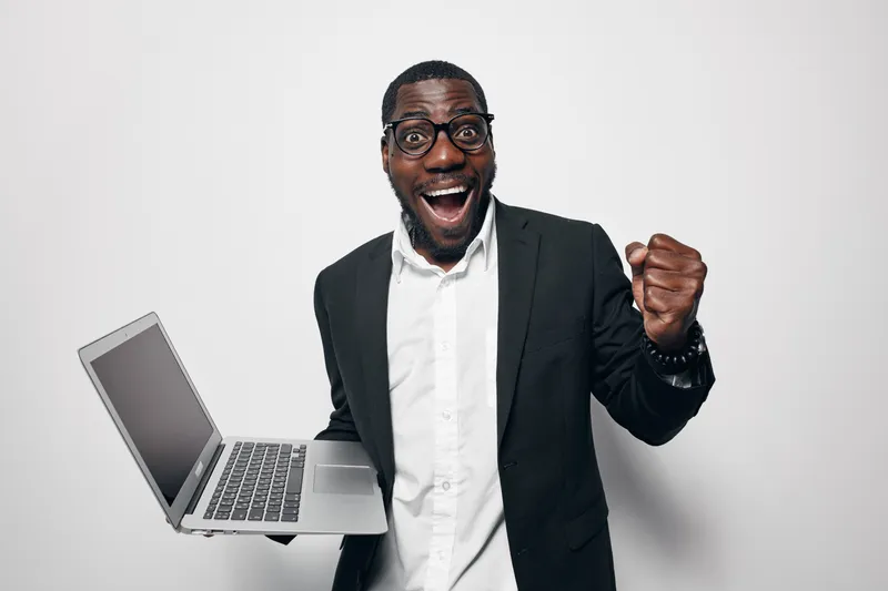Excited African American man with dark skin wearing glasses and black suit holding laptop celebrating success with fist pump on white background. Business, technology and achievement concept.