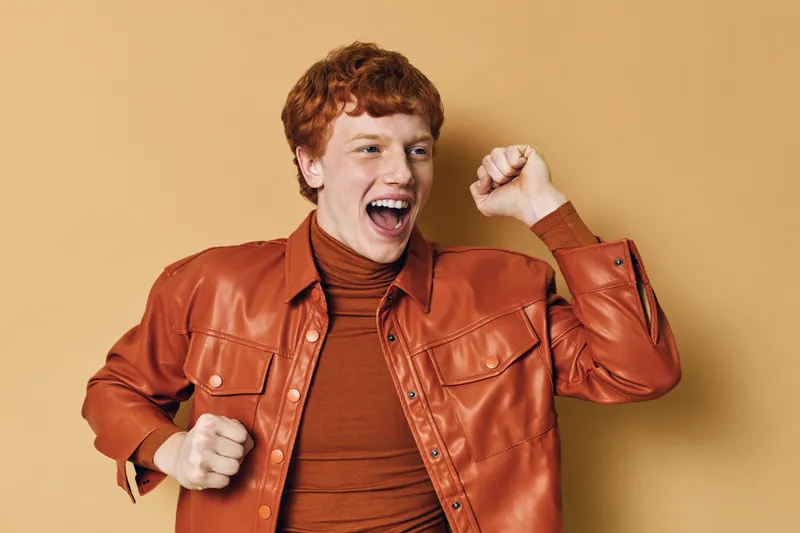 Redhead man wearing orange leather jacket and turtleneck celebrating with raised fist and open mouth against beige background, expressing happiness and excitement.