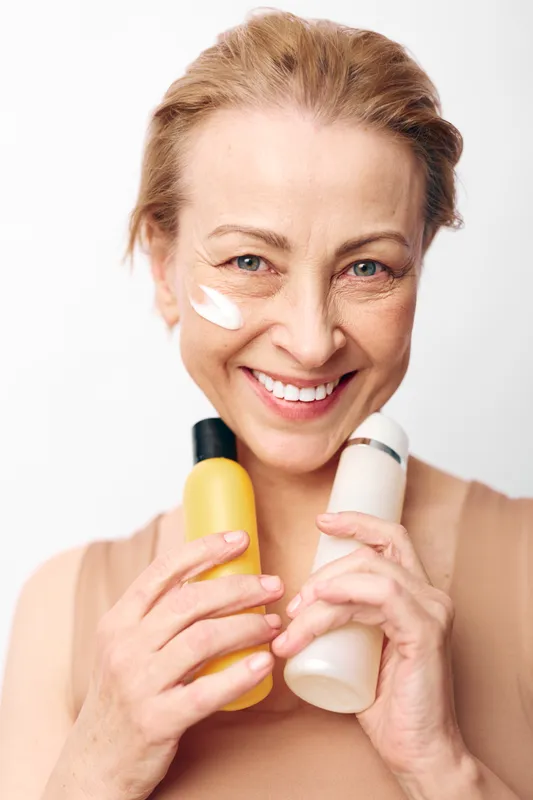 Smiling middle aged woman with short hair holds two cosmetic bottles close to her face with cream on cheek. Skincare routine concept, natural beauty, wellness, self care, healthy lifestyle.
