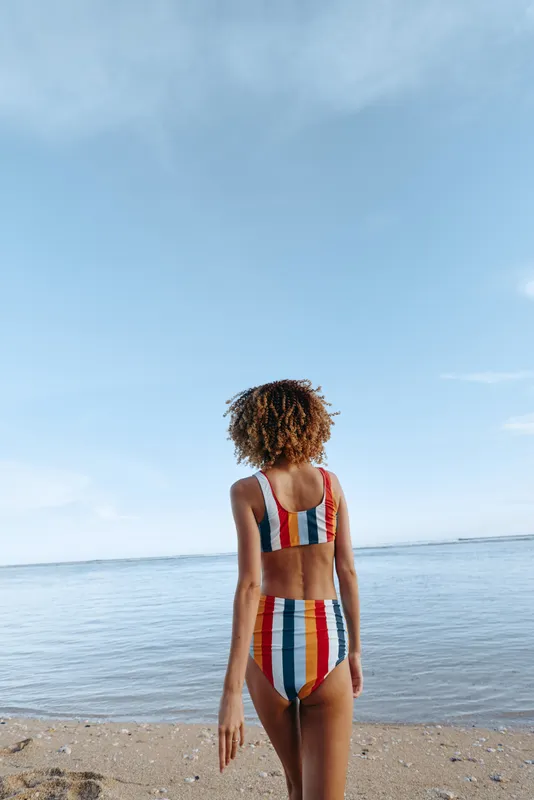 Woman on beach, ocean in background, striped swimsuit, summer travel mood, standing on sandy shoreline, she looks at blue sky, calm seaside horizon, solitude and relaxation