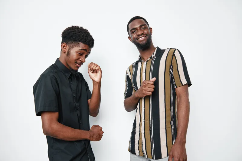 Two young black men with dark skin smiling and dancing together in casual clothes against white background. Friendship, joy, and positive energy in studio portrait.