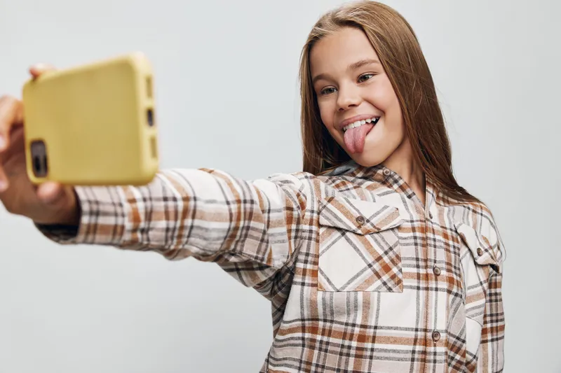 Teenage girl taking selfie with smartphone while smiling and sticking tongue out, wearing plaid shirt against plain background in casual lifestyle portrait photo.