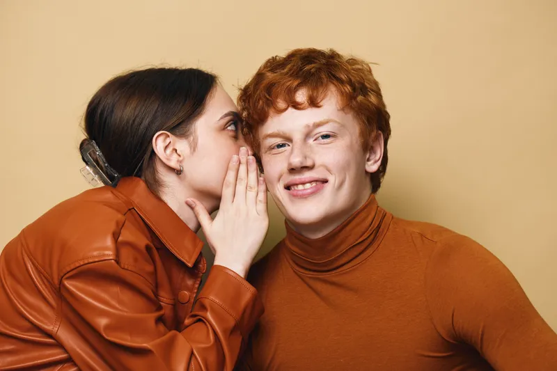 Young woman whispering secret to smiling young man with red hair in warm clothing on beige background. Closeup, communication, friendship, casual and cheerful emotions.