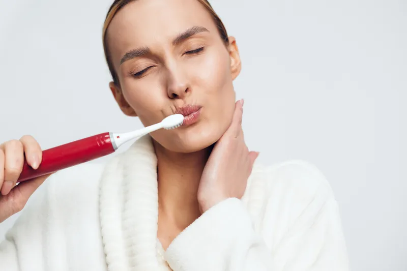 A young woman in a white bathrobe brushes her teeth with a red electric toothbrush, eyes closed, showing a relaxed morning routine in a bright minimalist setting.