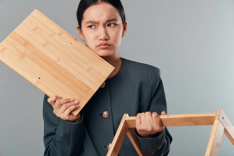 Young woman holding broken wooden furniture parts with frustrated expression on light background, standing indoors in casual clothes, repairing or assembling at home.