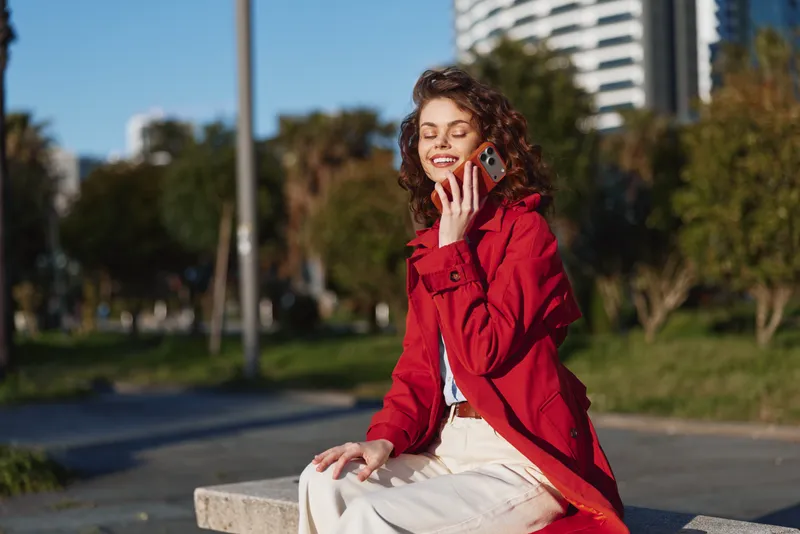 Cheerful woman outdoors in a red coat, smiling while talking on a smartphone, sunlit park scene with greenery and city background, relaxed pose with natural tones