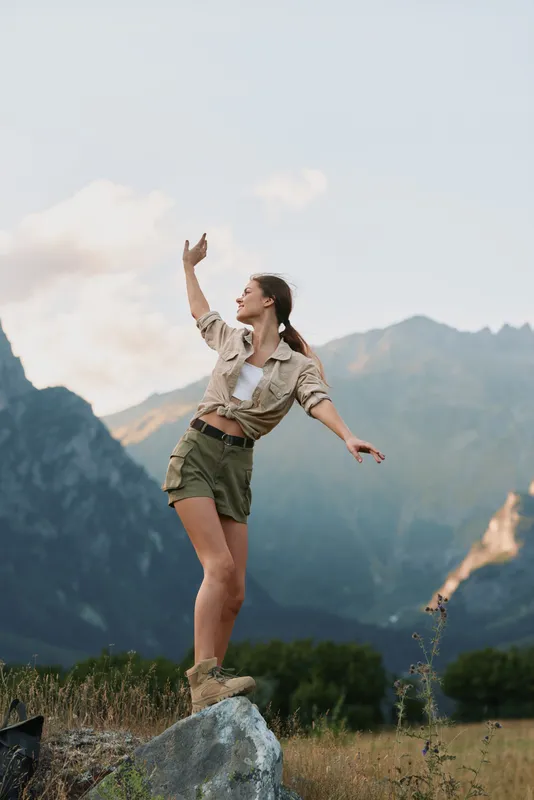 Young woman with brunette hair and ponytail balances on a rock in casual shorts and jacket against a mountain landscape during a clear day.