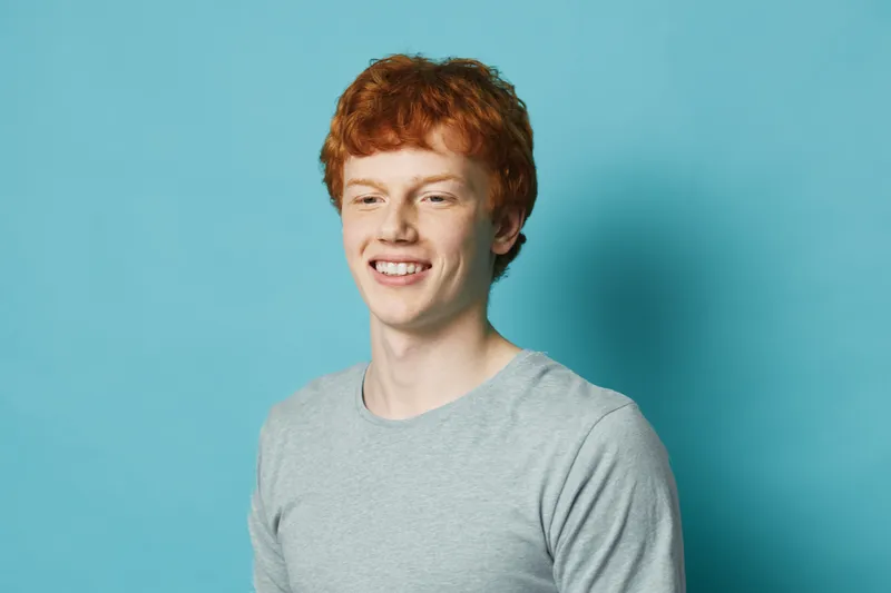 Young man with red hair smiling and wearing gray shirt standing against blue background. Portrait of happy male with casual look and cheerful expression in studio.