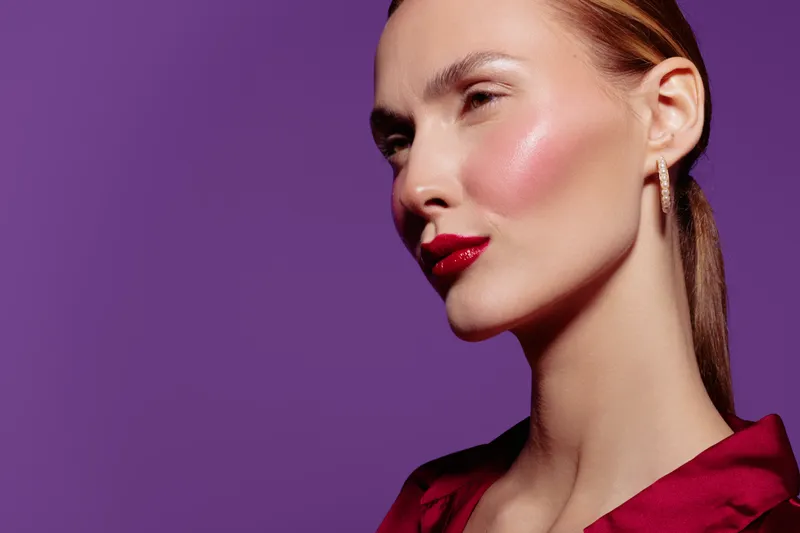 Close-up of a confident woman with red lipstick and glowing skin wearing red satin blouse against purple background. Concept of beauty, makeup, fashion, and elegance in studio lighting.